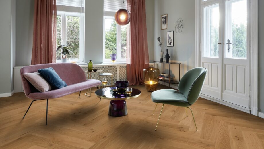 Helles, stilvolles Wohnzimmer mit Fischgrätparkett aus Eiche Mix (140 mm, geölt). Die Einrichtung kombiniert zartrosa Sofa, salbeigrünen Suunnittelustuhl und einen modernen Couchtisch mit goldenen Akzenten. Große weiße Flügeltüren und Fenster bringen viel Tageslicht in den Raum. Warme Vorhänge, moderne Kunstobjekte und eine elegante Ambienteleuchte schaffen eine harmonische und einladende Atmosphäre. Fischgrätparkett Eiche Mix 140 mm geölt im hellen Wohnzimmer mit pastellfarbenen Möbeln.