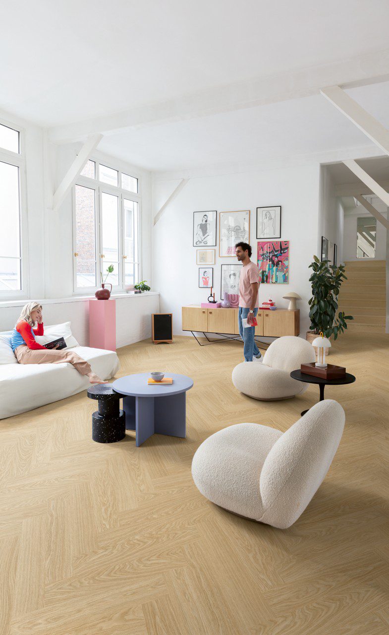 Klick Vinyl Fischgrat Quick Step Ciro Pure Oak Blush, verlegt auf dem Wohnzimmerboden und auf der Treppe mit farblich passender Incizo-Leiste. Eine Frau liegt auf einem weißen Sofa und unterhält sich mit einem Mann, der auf dem Boden steht.