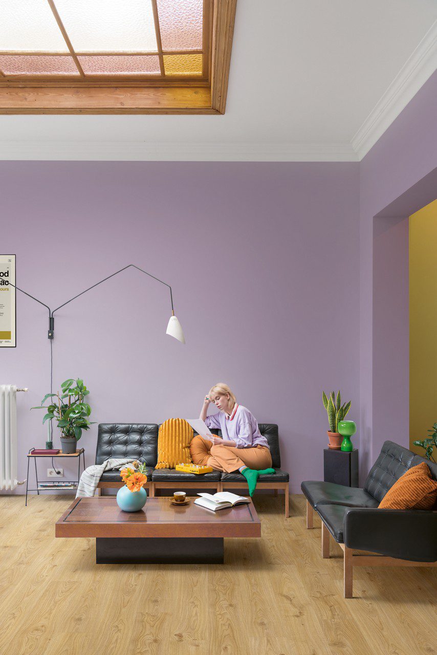 LVT parkett Quick-Step Blos Cottage tamm natural paigaldatud elutoa põrandale. Kaks nahast diivanit, diivanilaud lilledega. Naine istub diivanil ja loeb dokumenti.