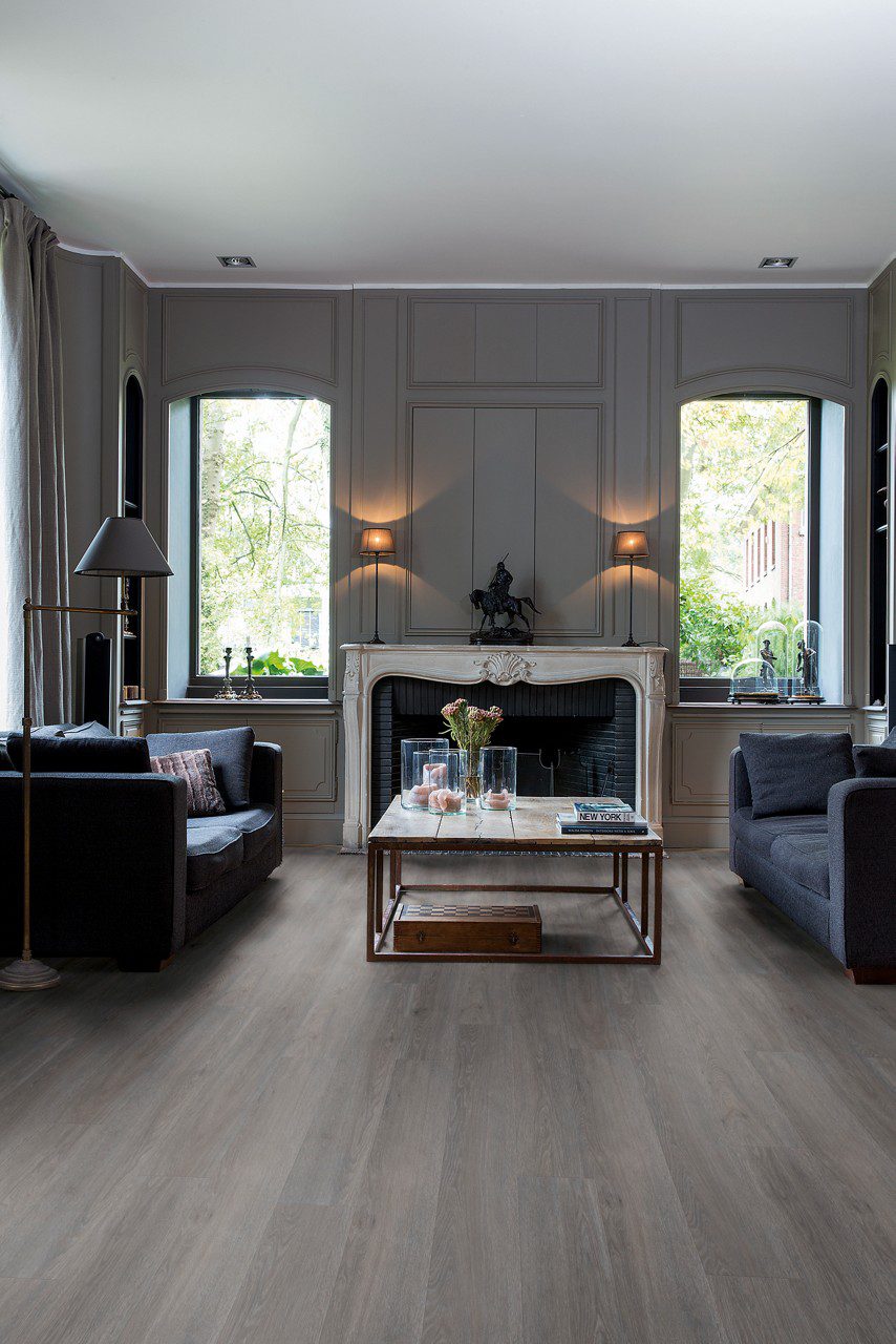 LVT parkett Quick-Step Blos Silk tamm dark grey paigaldatud klassikalise disainiga elutoa põrandale. Kaks diivanit suure diivanilaua ümber millel lilled ja küünland. Klassikalises stiilis valgest kivist kamin.