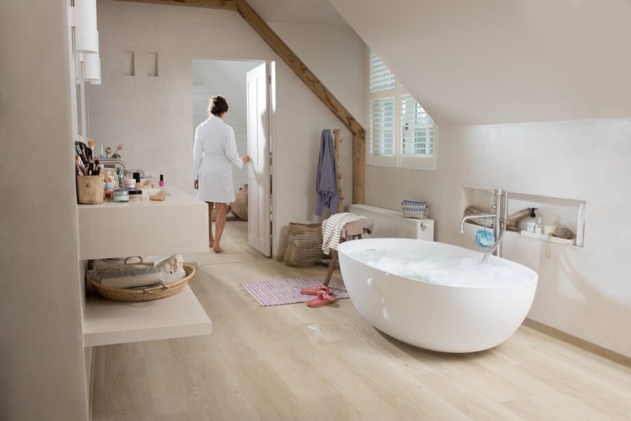 Vaalea vinyylilankku Quick Step Bloom Sea Breeze Oak Beige asennettuna kylpyhuoneen lattialle, erillinen valkoinen kylpyamme ja beige kylpyhuonekalusteet, nainen kävelee märin jaloin kylpytakissa lattialla.