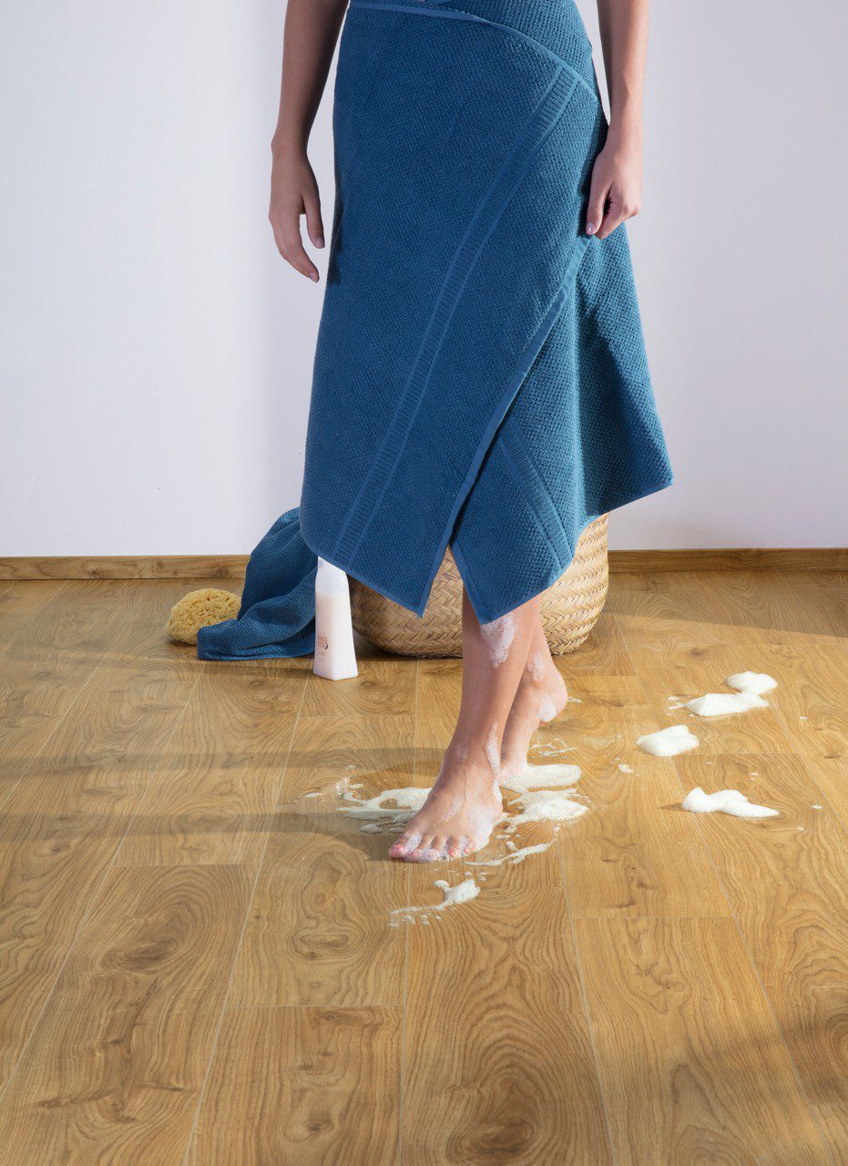 Vinyylilattia Quick Step Blos Cottage Tammi Natural asennettuna lattialle samansävyisten jalkalistojen kanssa, luonnollinen tammen sävy ja lankkulattia tammi, nainen kävelee lattialla märin ja vaahtoisin jaloin.