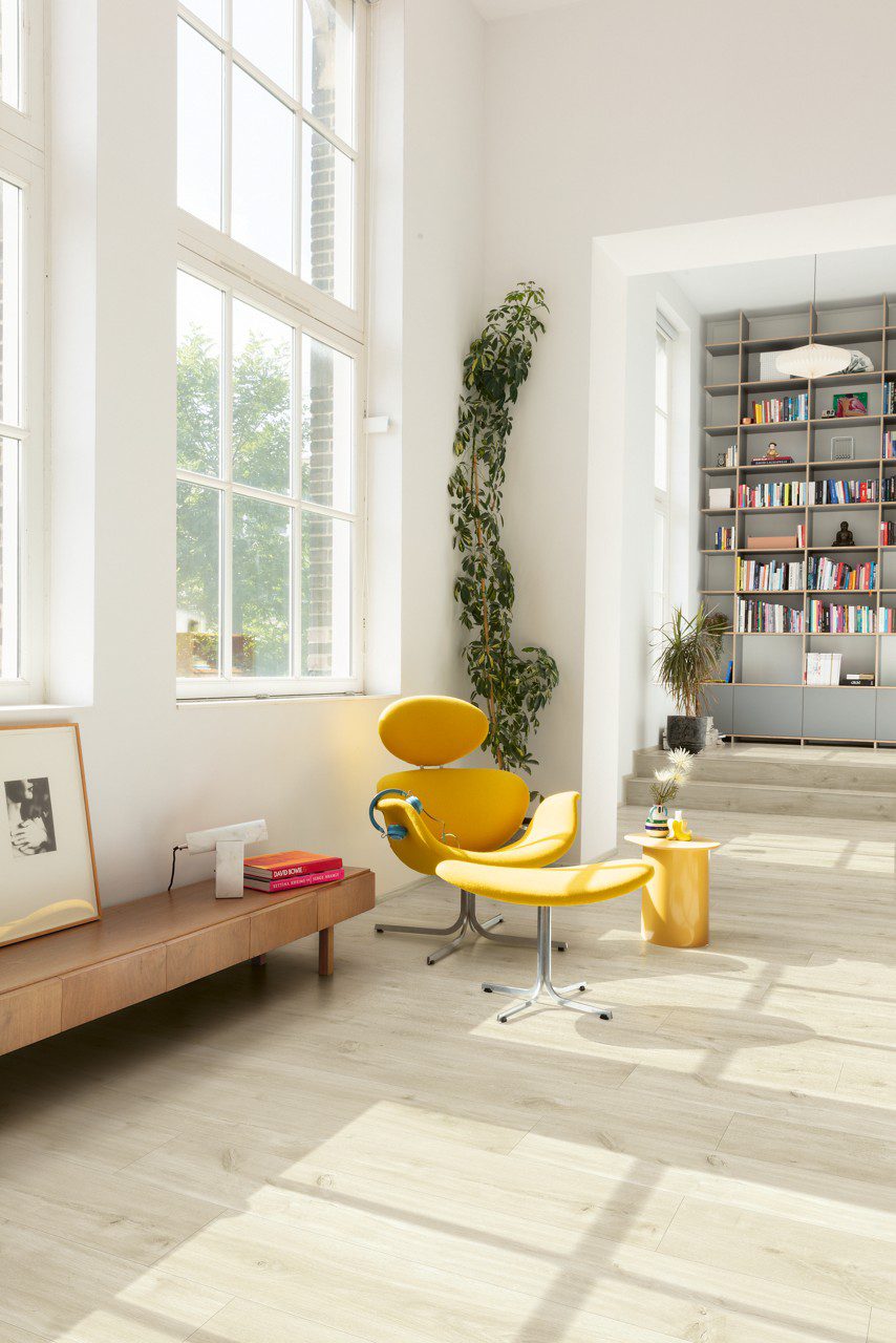 Vinyylilattia Quick Step Blos Vaalea Canyon Tammi Beige asennettuna olohuoneen lattialle ja kirjanurkkaan johtaville porrasaskelmille, vaalea hiekanbeige tammen sävy ja lankkudesign, keltainen lepotuoli jalkarahilla, lipasto sekä hyllyjä kirjoineen.