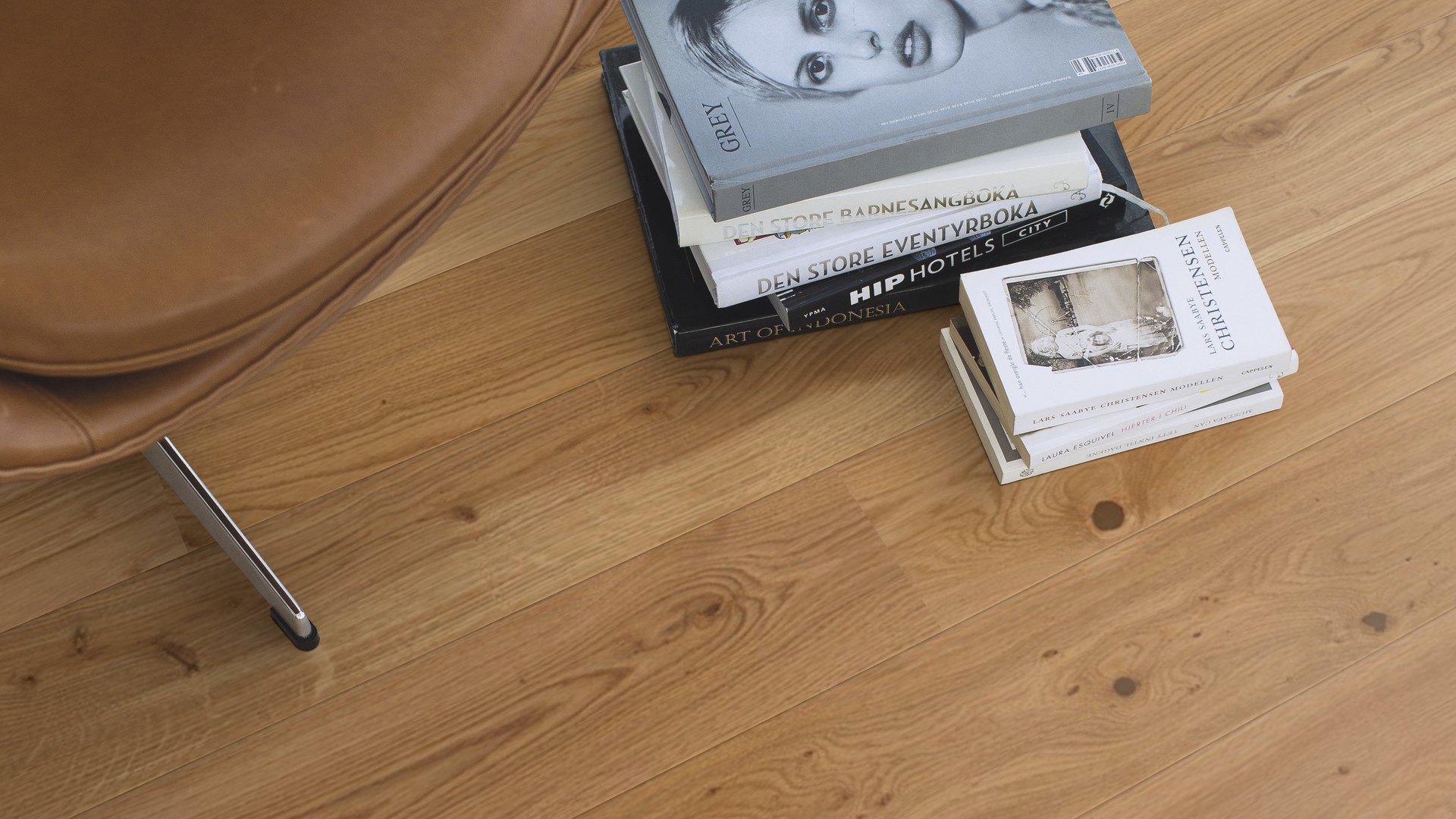 Tammiparketti Bauwerk Boen Casapark tammi 46 Vivo (138 mm), harjattu ja öljyvahattu, asennettuna olohuoneen lattialle – vaaleanruskealla nahalla verhoiltu nojatuoli ja kirjoja lattialla. Luonnollinen Vivo-sävy ja rustiikkinen sorteerus korostavat tammen eläväistä kuviointia.