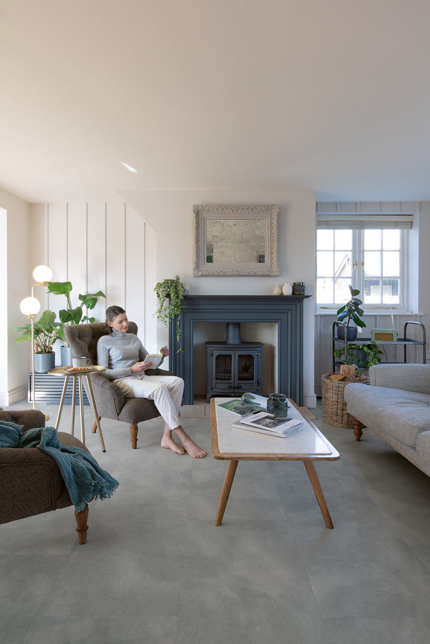 Quick-Step Illume Soft Sky vinyl floor tiles installed on a living room floor – a woman reads a book in a comfortable armchair; a sofa, table and a classic metal fireplace in the room. Light, fresh grey-blue tone with a subtle stone-inspired design.