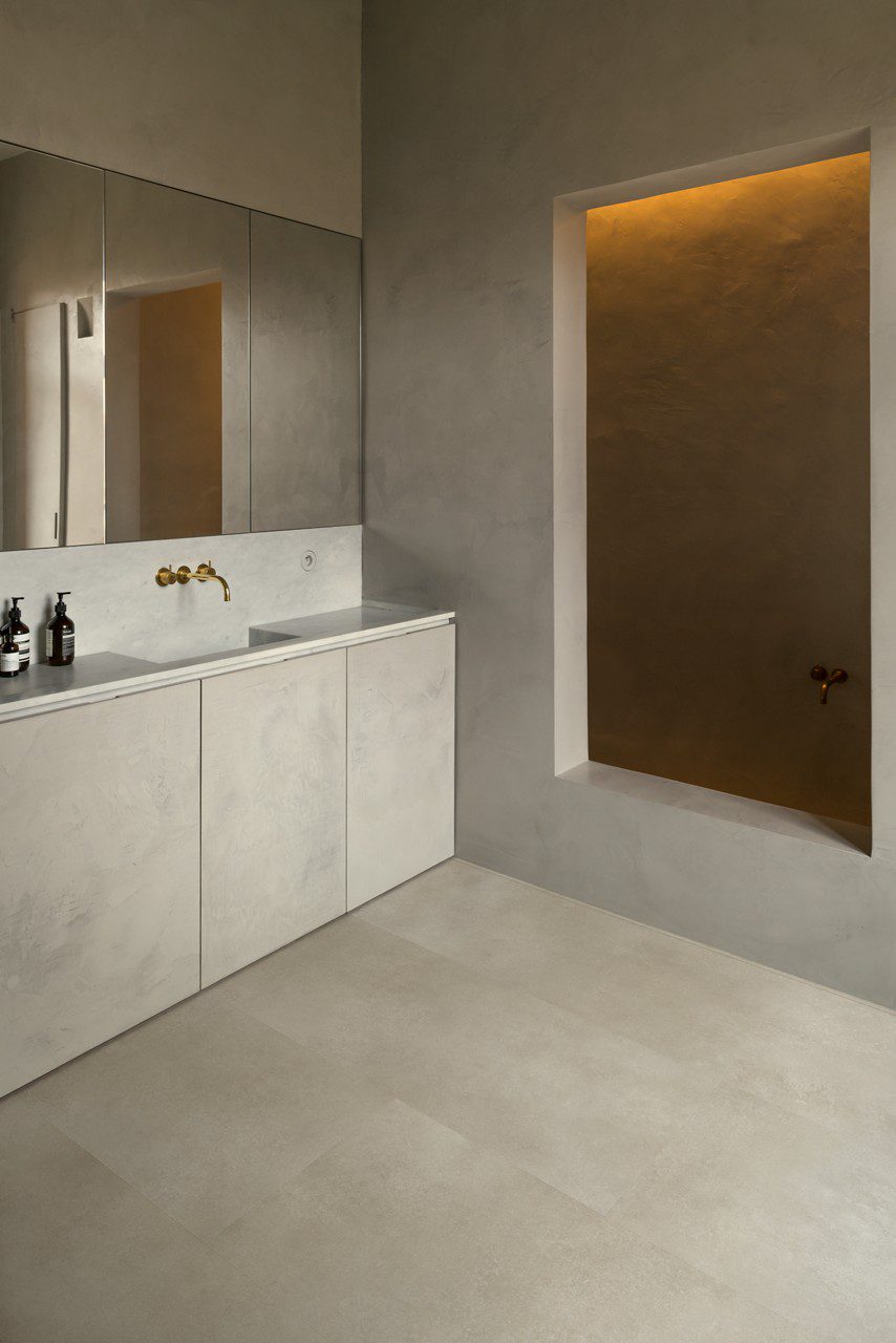 Vinylboden in Betonoptik Quick-Step Illume Sandstone Concrete, verlegt auf dem Badezimmerboden – graues Badmöbel mit grauer Waschtischplatte und Aufsatzbecken, drei Spiegelschränke an der Wand sowie eine goldene Wandarmatur; warmer Greige-/Sandton mit dezentem Betondesign.