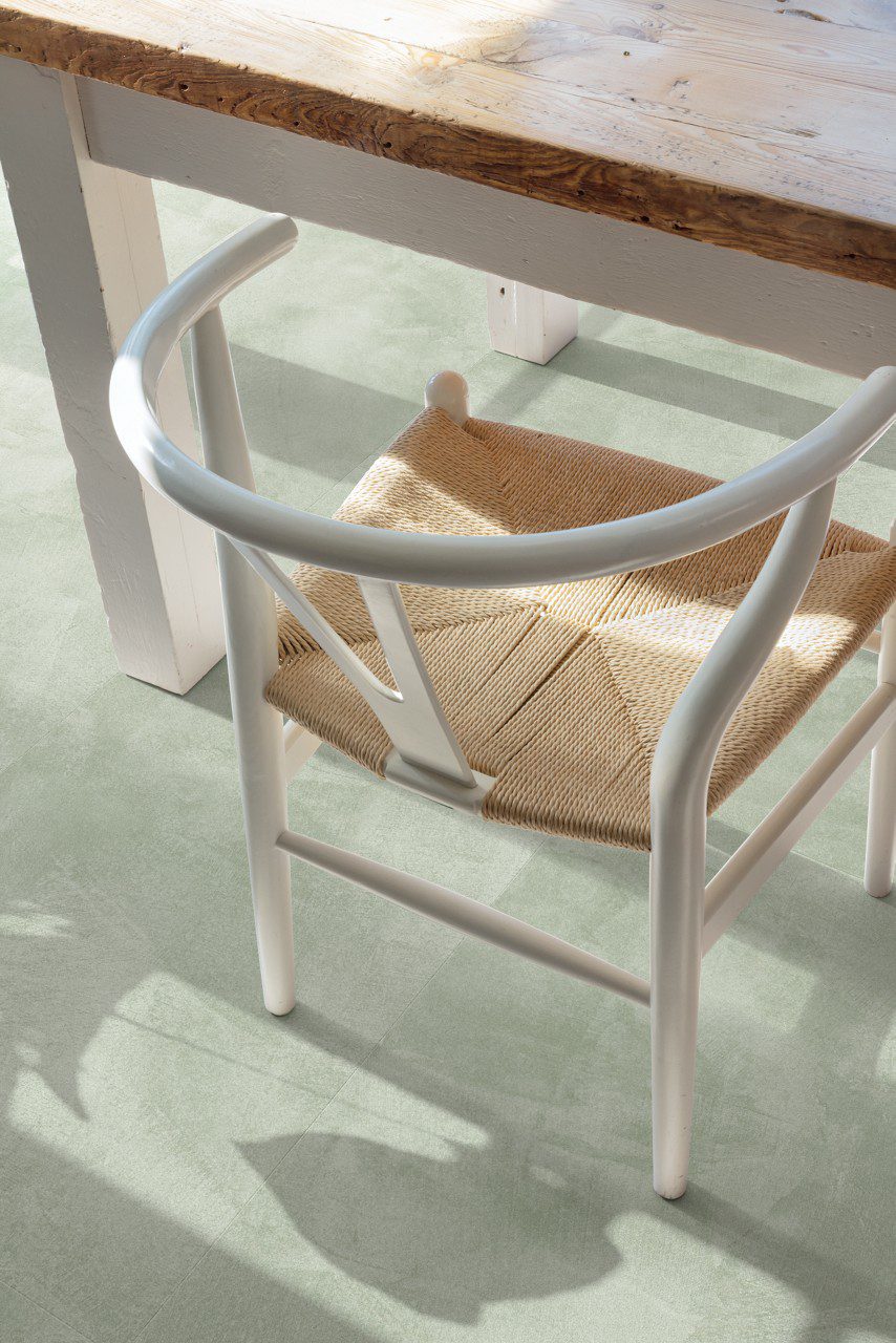Vinylboden in Betonoptik Quick-Step Illume Soft Sage, verlegt auf dem Küchenboden – rustikaler Esstisch und Stuhl in Nahaufnahme; heller, sanfter Salbeigrün-Ton mit dezentem, steinartigem Suunnittelu.