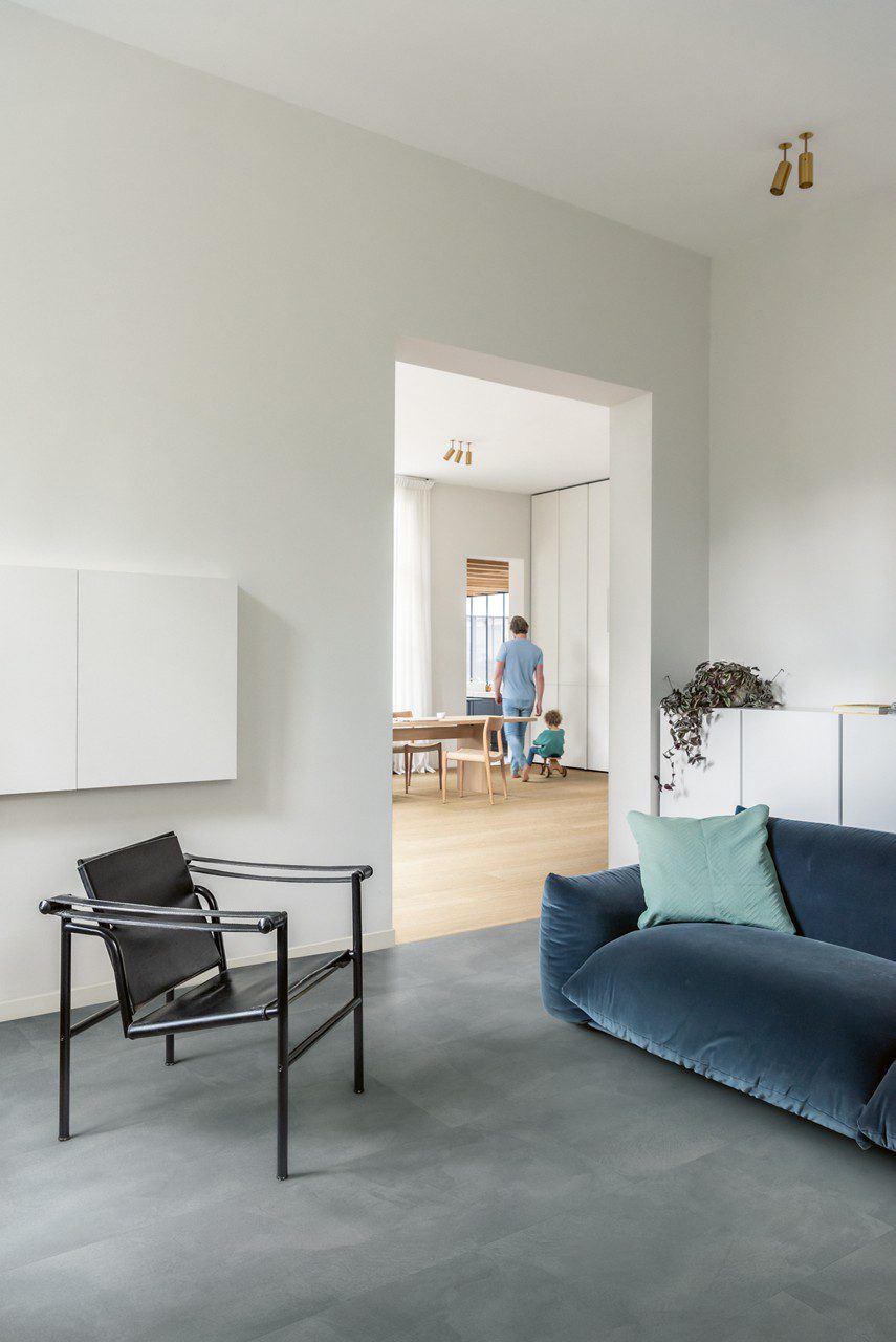 Vinylboden in Betonoptik Quick-Step Illume Soft Sky, verlegt auf dem Wohnzimmerboden – schwarzer Ledersessel und blaues Sofa; im Hintergrund sind in der Küche ein stehender Vater und ein kleiner Sohn auf einem Dreirad zu sehen.