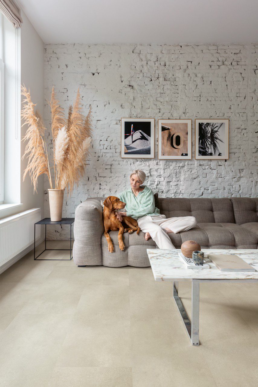 Vinylboden in Betonoptik, verlegt auf dem Wohnzimmerboden – weiß gestrichene rustikale Ziegelwand, Couchtisch aus weißem Marmor mit verchromten Metallbeinen und graues Sofa; eine blonde Frau streichelt ihren braunen Hund.
