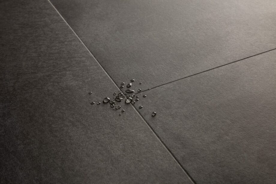 vinyylilattia-quick-step-illume-soft-graphite Vinyylilattia Quick-Step Illume Soft Graphite asennettuna kodin lattialle – tumma grafiitinharmaa sävy ja hillitty kivimäinen/betonimainen design hienovaraisella tekstuurilla; lattian pinnalla näkyy vesipisaroita korostamassa sen vedenkestävyyttä.