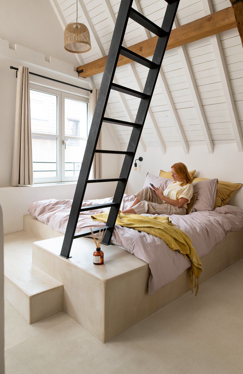 Кварц-винил Quick-Step Illume Sandstone Concrete уложен на полу мансардной спальни — женщина лежит в постели и читает книгу; тёплый песочно-бежевый (greige) оттенок и спокойный бетонный дизайн.