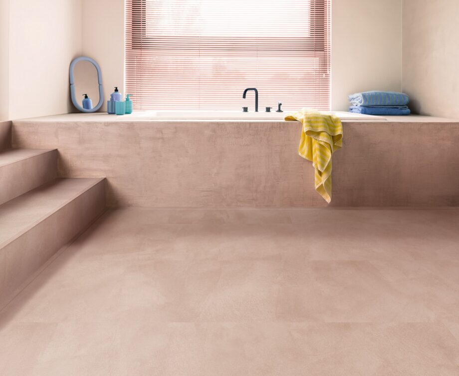 кварц-винил-quick-step-illume-soft-blush Кварц-винил Quick-Step Illume Soft Blush уложен на полу ванной комнаты и на ступенях лестницы — белая ванна встроена в конструкцию в розовом тоне, совпадающем по оттенку с полом; светлый нежно-розово-бежевый оттенок и спокойный каменно-бетонный дизайн.