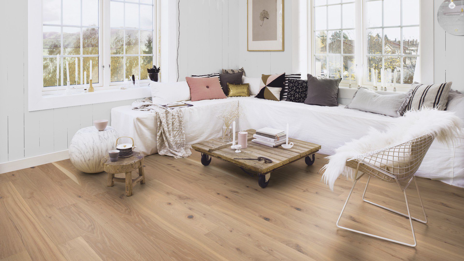 Паркетная доска Bauwerk Boen Casapark дуб 46 Vivo (138 мм), брашированная, масло-воск — уложена на полу гостиной в рустикальном стиле; белый угловой диван с подушками разных цветов и низкий журнальный столик на колёсиках из деревянных брусьев. Рустикальная сортировка с выразной текстурой и сучками.