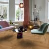 Helles, stilvolles Wohnzimmer mit Fischgrätparkett aus Eiche Mix (140 mm, geölt). Die Einrichtung kombiniert zartrosa Sofa, salbeigrünen Suunnittelustuhl und einen modernen Couchtisch mit goldenen Akzenten. Große weiße Flügeltüren und Fenster bringen viel Tageslicht in den Raum. Warme Vorhänge, moderne Kunstobjekte und eine elegante Ambienteleuchte schaffen eine harmonische und einladende Atmosphäre. Fischgrätparkett Eiche Mix 140 mm geölt im hellen Wohnzimmer mit pastellfarbenen Möbeln.