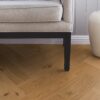 kalanruotoparketti-tammi-mix-140mm-4-sivulta-viistetty-harjattu-osmo-oljyvahaa Kalanruotoparketti Tammi Mix 140 mm, 4 sivulta viistetty, harjattu, Osmo öljyvahaa – asennettu eteisen lattialle. Beigenvärinen sohva mustilla jaloilla, valkoiseksi maalatut jalkalistat, harmaat seinäpaneelit, valkoinen roskakori.