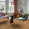 Helles, stilvolles Wohnzimmer mit Fischgrätparkett aus Eiche Mix (140 mm, geölt). Die Einrichtung kombiniert zartrosa Sofa, salbeigrünen Suunnittelustuhl und einen modernen Couchtisch mit goldenen Akzenten. Große weiße Flügeltüren und Fenster bringen viel Tageslicht in den Raum. Warme Vorhänge, moderne Kunstobjekte und eine elegante Ambienteleuchte schaffen eine harmonische und einladende Atmosphäre. Kalanruotoparketti Tammi Mix öljyvahattu 140mm – 9