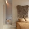 klickvinyl-quick-step-bloom-sea-breeze-oak-light Klickvinyl Quick Step Bloom Sea Breeze Oak Light im Schlafzimmer verlegt, sehr heller Eicheton und Dielendesign, Bett mit schöner hellbrauner Bettwäsche, im Hintergrund ein Badezimmer mit freistehender, eckiger Suunnittelu-Badewanne.
