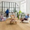LVT floors Quick-Step Bloom Elegant Oak Natural installed on a living room floor. Woman is sitting on a chair and reading a book. Man is looking the vinyl record player. Table with books, palm threes