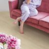 Vinyl flooring Quick-Step Bloom Sea Breeze Oak Beige installed on living room floor. Woman sitting barefoot on a pink sofa and drinking cocktail. Pink flowers.
