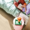 Vinylboden Quick Step Bloom Brushed Oak Beige im Schlafzimmer, Frau liest ein Buch auf dem Bett, rosa Sitzhocker.