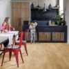 Vinyylilattia Quick Step Blos Canyon Tammi Natural asennettuna ruokailutilan lattialle, luonnollinen tammen sävy ja lankkulattia tammi, äiti istuu ruokapöydän ääressä ja seuraa tytärtä, joka ottaa banaanin hedelmävadilta keittiön kaapista.