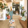 Vinyylilattia Quick Step Blos Coast Tammi Beige asennettuna olohuoneen lattialle ja huoneita erottavalle matalalle portaalle, vaalea hiekanbeige tammen sävy ja lankkulattia tammi, nainen kävelee portaita ylös ja koira odottaa häntä.