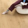 Виниловый ламинат Quick Step Bloom Elegant Oak Natural уложен на полу в гостиной, женщина сидит в удобном кресле и читает книгу.