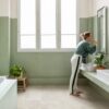 кварц-винил-quick-step-illume-oyster-concrete Кварц-винил Quick-Step Illume Oyster Concrete уложен на полу ванной комнаты — мебель с двумя накладными раковинами; женщина опирается рукой на раковину и чистит зубы, глядя на своё отражение в настенном зеркале.