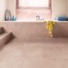 кварц-винил-quick-step-illume-soft-blush Кварц-винил Quick-Step Illume Soft Blush уложен на полу ванной комнаты и на ступенях лестницы — белая ванна встроена в конструкцию в розовом тоне, совпадающем по оттенку с полом; светлый нежно-розово-бежевый оттенок и спокойный каменно-бетонный дизайн.